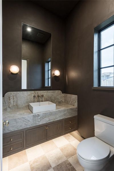 This powder bathroom is an intimate study in drama and materiality. Venetian plaster walls in a deep, moody tone create a cocoon-like effect, while a striking stone vanity with seamless waterfall edges adds sculptural weight. The vessel sink and wall-mounted fixtures bring a touch of modernity, while alabaster sconces offer a soft, ambient glow. Under-cabinet lighting washes over the checkerboard stone floors, tying the room together with a quiet elegance that’s anything but ordinary. This powder bathroom is an intimate study in drama and materiality. Venetian plaster walls in a deep, moody tone create a cocoon-like effect, while a striking stone vanity with seamless waterfall edges adds sculptural weight. The vessel sink and wall-mounted fixtures bring a touch of modernity, while alabaster sconces offer a soft, ambient glow. Under-cabinet lighting washes over the checkerboard stone floors, tying the room together with a quiet elegance that’s anything but ordinary.