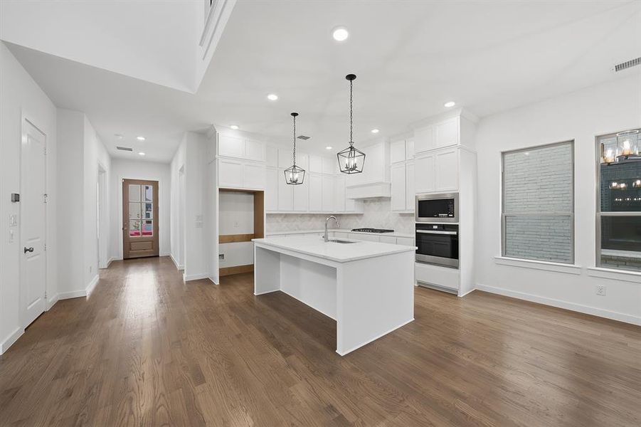 Kitchen featuring white cabinets, appliances with stainless steel finishes, a center island with sink, tasteful backsplash, and hanging light fixtures