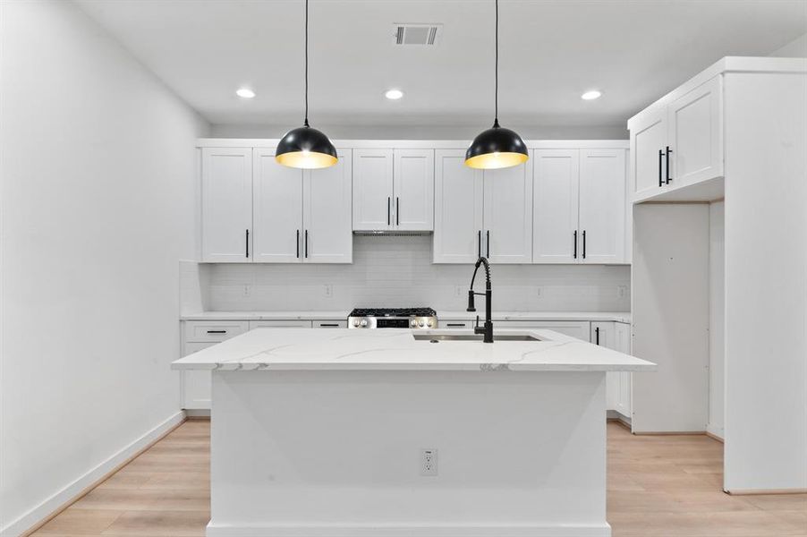 Modern kitchen with a sleek, minimalist design. Features white cabinetry, a central island with a sink, black pendant lighting, and hardwood floors.