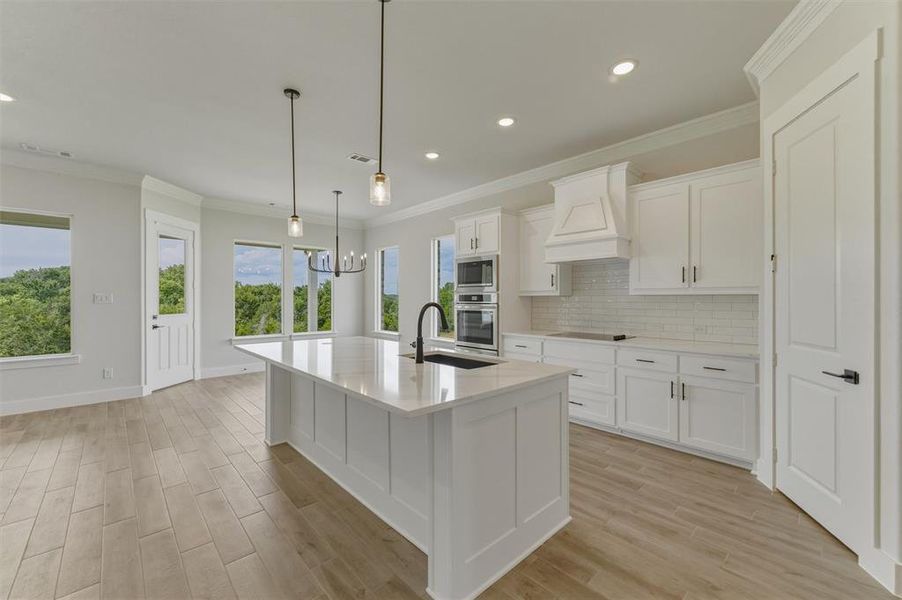 Open-concept kitchen featuring an oversized island with a built-in sink, white shaker cabinetry, light-toned wood-finish flooring, and a subway tile backsplash Open-concept kitchen featuring an oversized island with a built-in sink, white shaker cabinetry, light-toned wood-finish flooring, and a subway tile backsplash