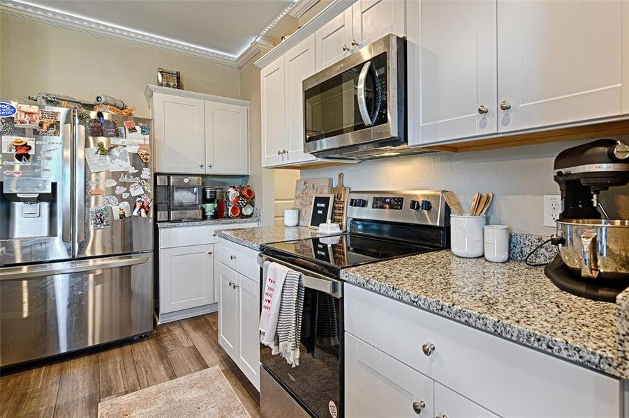 Kitchen featuring appliances with stainless steel finishes, white cabinets, ornamental molding, light wood-style floors, and light stone counters Kitchen featuring appliances with stainless steel finishes, white cabinets, ornamental molding, light wood-style floors, and light stone counters