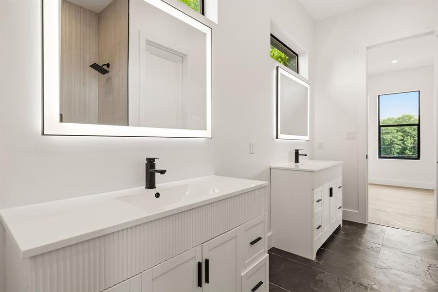 Dual vanity bathroom featuring backlit mirrors, matte black fixtures, crisp white cabinetry with fluted details, and dark tile flooring