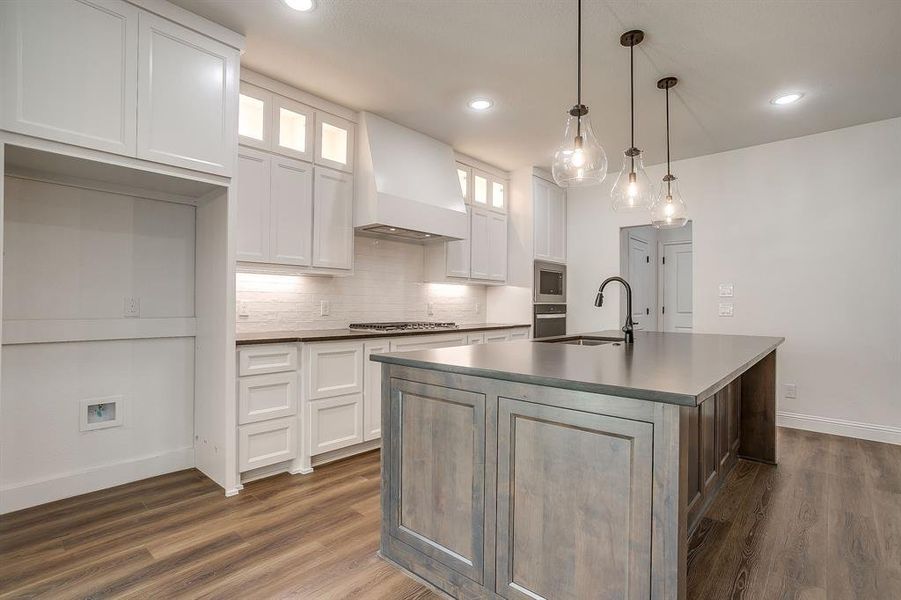 Kitchen with glass insert cabinets, dark countertops, white cabinets, an island with sink, and recessed lighting
