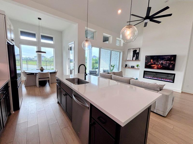 The open-concept living space features a sleek kitchen with a large island, a cozy living area with a mounted TV and fireplace, and abundant natural light from large windows and sliding glass doors leading to an outdoor view.