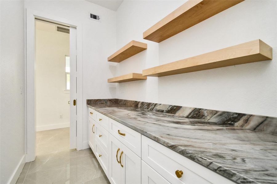 High-end prep space featuring premium stone countertops, white cabinetry with gold hardware, and floating natural wood shelving.