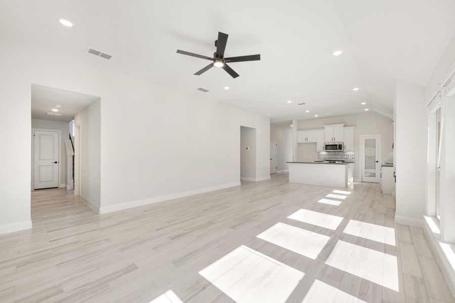 Open living area with light wood floors, ceiling fan, and white kitchen with island in background