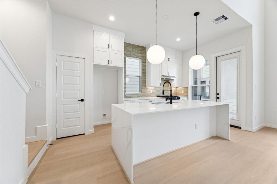 Large waterfall-edge island anchors the kitchen, offering generous workspace and seating. Matte black fixtures and quartz surfaces create a polished, modern feel that flows into the open-concept living area with soaring 20-ft ceilings. This shot also highlights the door leading out to the balcony.