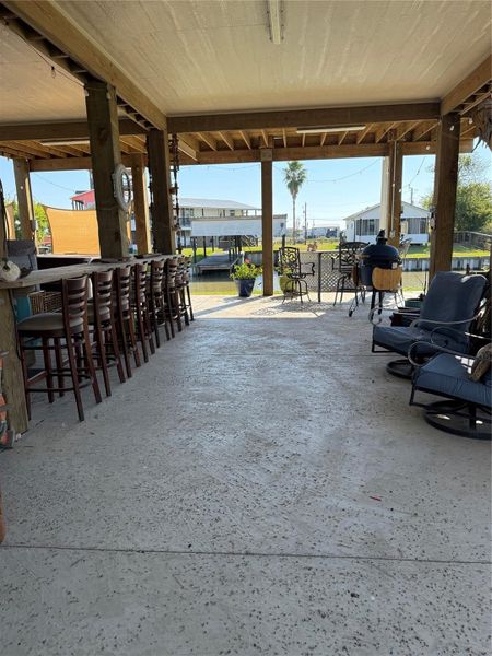Spacious area under house with bar seating and cozy outdoor furniture, offering a relaxing view of the canal and nearby homes. Perfect for entertaining and enjoying waterfront living.