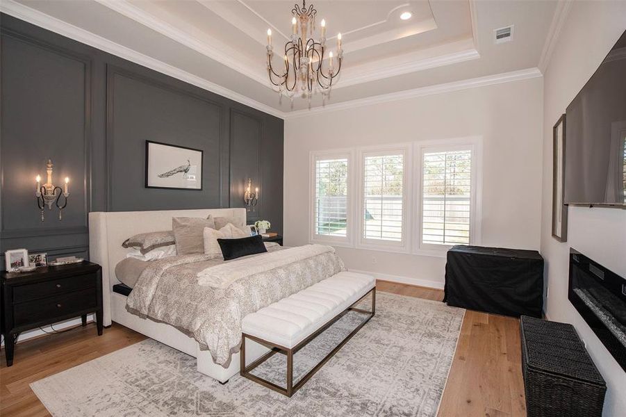 This bedroom features elegant tray ceilings with a chandelier, a cozy upholstered bed, and a stylish dark accent wall with sconces. Large windows with shutters provide natural light, while a modern fireplace and hardwood floors add warmth and luxury.