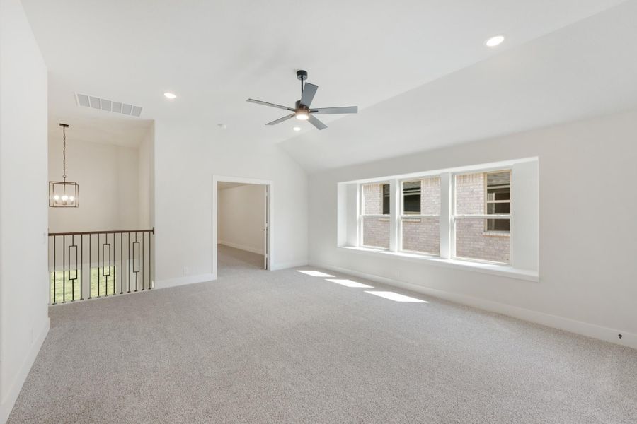 Spacious empty room with vaulted ceiling, ceiling fan, carpet flooring, and large windows near an open staircase landing.