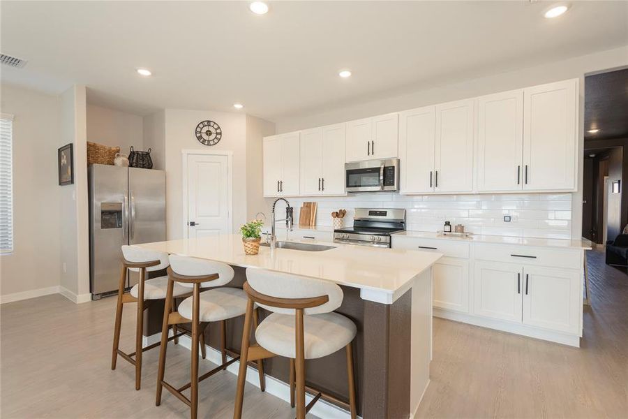 Modern kitchen featuring a central island with countertop seating, white shaker-style cabinetry, stainless steel appliances, a subway tile backsplash, and light wood-finish flooring Modern kitchen featuring a central island with countertop seating, white shaker-style cabinetry, stainless steel appliances, a subway tile backsplash, and light wood-finish flooring