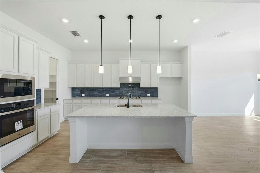 Spacious kitchen featuring an expansive center island with pendant lighting, stainless steel built-in appliances, white shaker cabinetry, and a dark tile backsplash