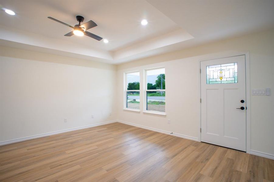 Open living area featuring wood-finish flooring, a tray ceiling with recessed lighting and a ceiling fan, and a white entry door with stained glass insert
