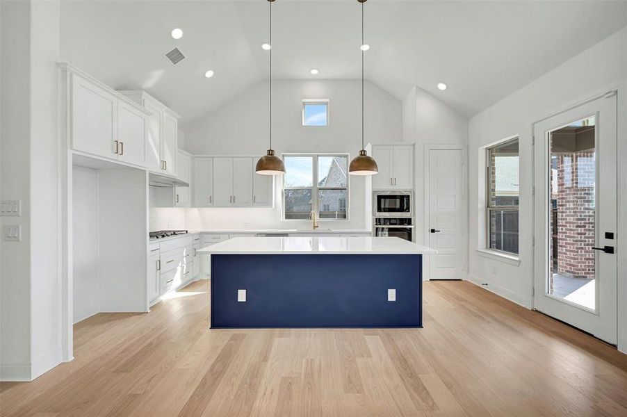 Kitchen with white cabinetry, a kitchen island, a high ceiling, hanging light fixtures, and light wood finished floors Kitchen with white cabinetry, a kitchen island, a high ceiling, hanging light fixtures, and light wood finished floors