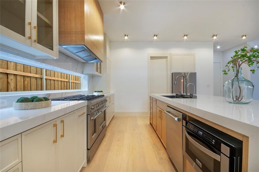 Kitchen with light stone counters, appliances with stainless steel finishes, light wood-style flooring, glass insert cabinets, and a center island with sink Kitchen with light stone counters, appliances with stainless steel finishes, light wood-style flooring, glass insert cabinets, and a center island with sink