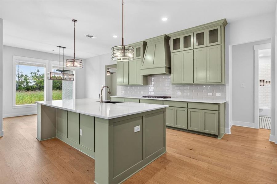 Spacious kitchen featuring a large island with a sink, light green cabinetry, white countertops, subway tile backsplash, and light wood-finish flooring Spacious kitchen featuring a large island with a sink, light green cabinetry, white countertops, subway tile backsplash, and light wood-finish flooring