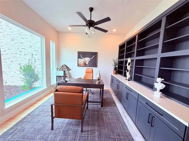 Dedicated study featuring expansive windows, wood-finish flooring, a contemporary ceiling fan, built-in dark cabinetry with integrated shelving, and a light-toned countertop