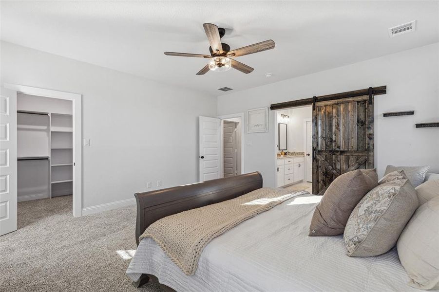 Carpeted bedroom featuring a barn door, ceiling fan, a walk in closet, a closet, and ensuite bath