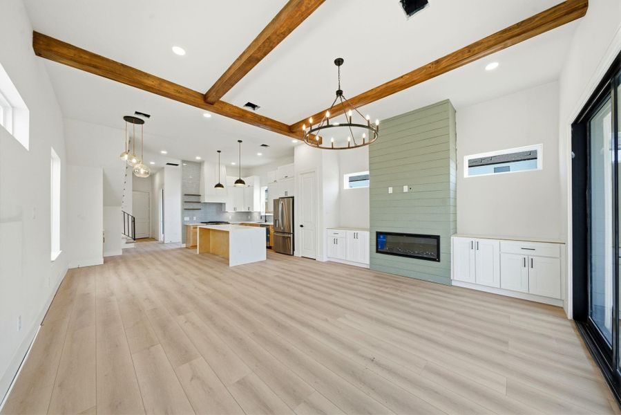 This spacious open-concept living area features light wood flooring, exposed wooden beams, and a modern kitchen with an island. A stylish chandelier and linear fireplace add elegance, while large windows and sliding doors provide ample natural light.