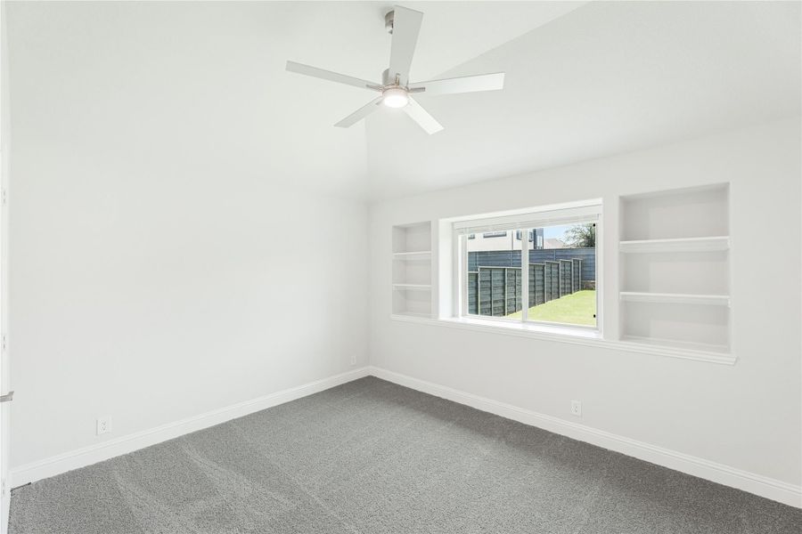 Empty bedroom with gray carpet, ceiling fan, built-in shelves, and a window overlooking a fenced backyard Empty bedroom with gray carpet, ceiling fan, built-in shelves, and a window overlooking a fenced backyard