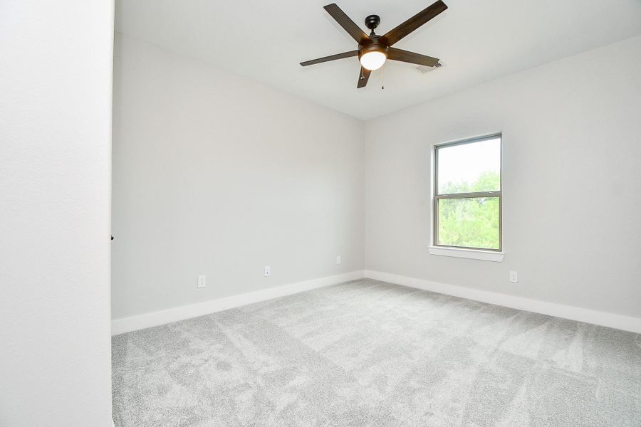 This is a bright, 4th bedroom featuring light gray walls, a textured gray carpet, and a ceiling fan with a light fixture. It has a single window providing natural light and a view of greenery. This is a bright, 4th bedroom featuring light gray walls, a textured gray carpet, and a ceiling fan with a light fixture. It has a single window providing natural light and a view of greenery.