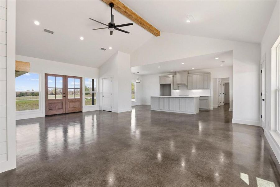 This expansive open-concept living area features polished concrete floors, a vaulted ceiling with a substantial wooden beam, and a ceiling fan