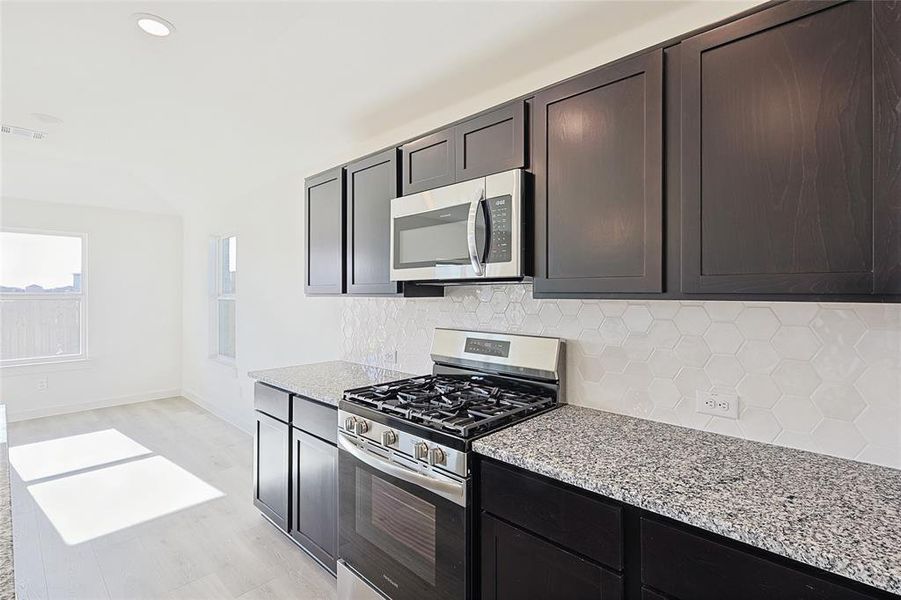 Kitchen with decorative backsplash, light stone countertops, appliances with stainless steel finishes, light hardwood / wood-style floors, and dark brown cabinetry