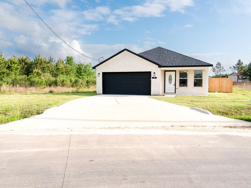 Charming single-story home with a modern exterior featuring a dark garage door and trim. It has a spacious driveway and is set against a backdrop of greenery, offering a serene and private setting.
