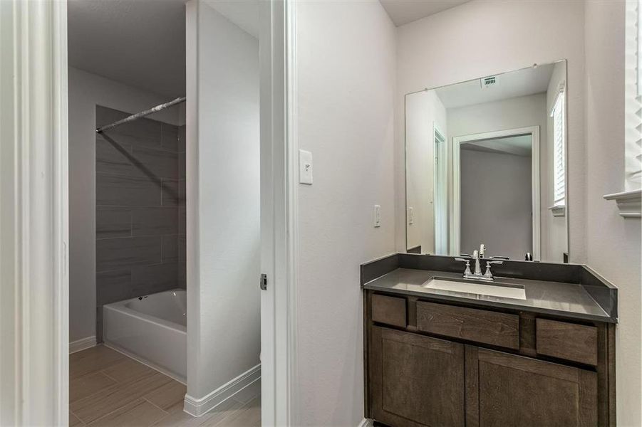 This modern bathroom vanity features sleek dark cabinetry complemented by a glossy countertop, offering ample space for all your essentials. The large mirror and elegant fixtures enhance the room's contemporary appeal, while the adjacent shower area, visible through the doorway, boasts a stylish tiled finish for a cohesive look. Photos are from another Rylan floor plan.