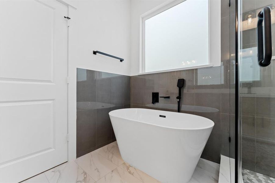 This modern bathroom features a sleek freestanding bathtub, elegant gray tile accents, and a glass shower enclosure. A large frosted window provides privacy while allowing natural light to brighten the space.