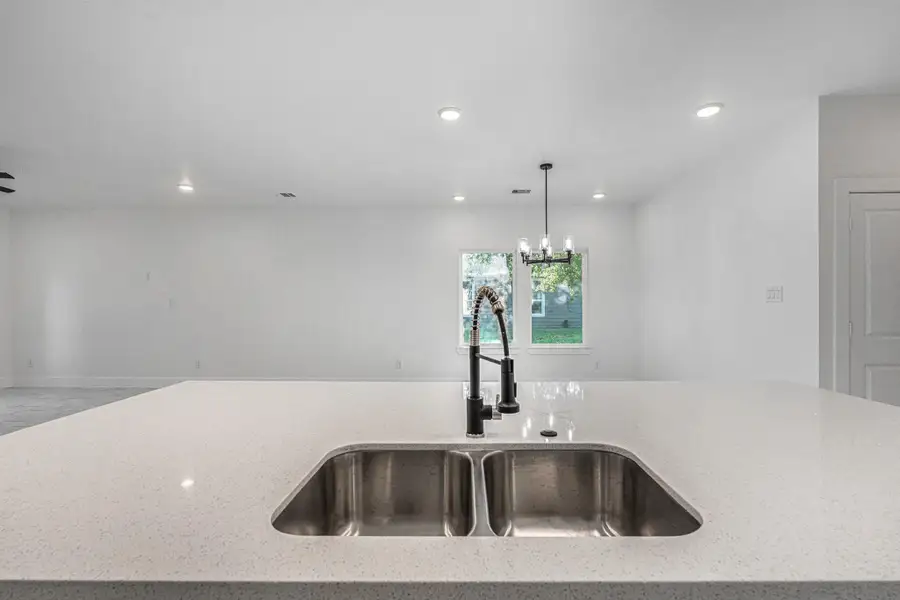 Modern, open-concept kitchen with a sleek countertop featuring a double sink and contemporary faucet. The space is bright with recessed lighting and a stylish chandelier, offering a view of the living area and outdoor greenery.