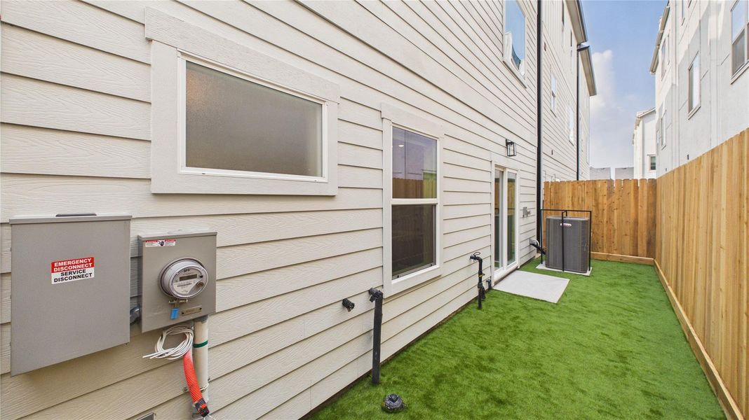 This photo shows a narrow side yard with artificial grass, a wooden fence, and access to the home's utilities. The siding is light-colored, and there are multiple windows and a patio door leading to the area.