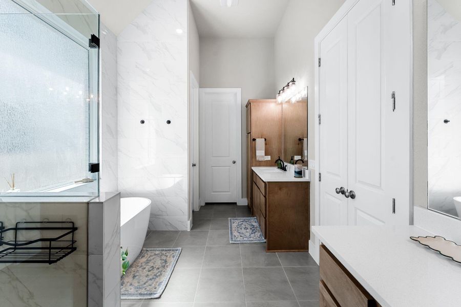Spacious bathroom featuring a frameless glass shower enclosure, a freestanding soaking tub, wood-finish double vanity with white countertops, and expansive tile flooring