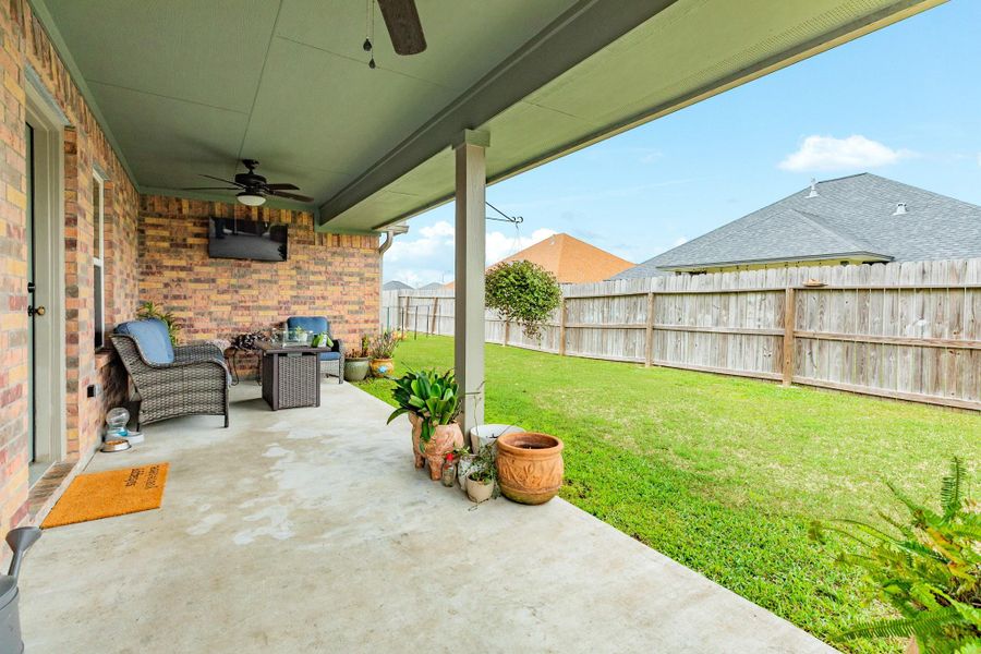 This photo showcases a covered patio with cozy seating and a ceiling fan, overlooking a well-maintained fenced backyard. Perfect for relaxing or entertaining outdoors.