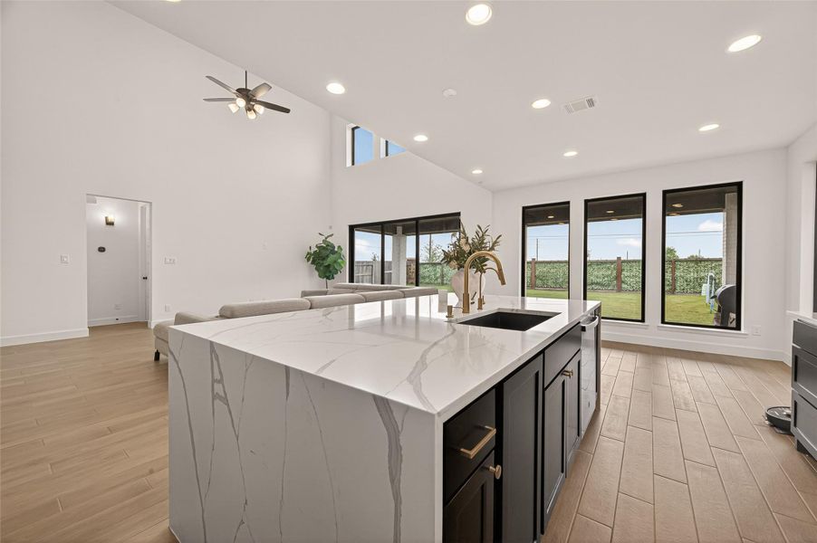 This modern open-concept living space features a spacious kitchen island with sleek countertops, recessed lighting, and large windows offering plenty of natural light. The room has high ceilings, wood-look flooring, and an inviting view of the backyard.