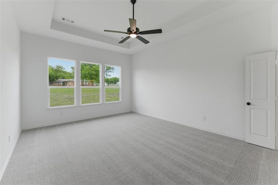 This spacious room features light gray carpeting, a tray ceiling, and a ceiling fan with light