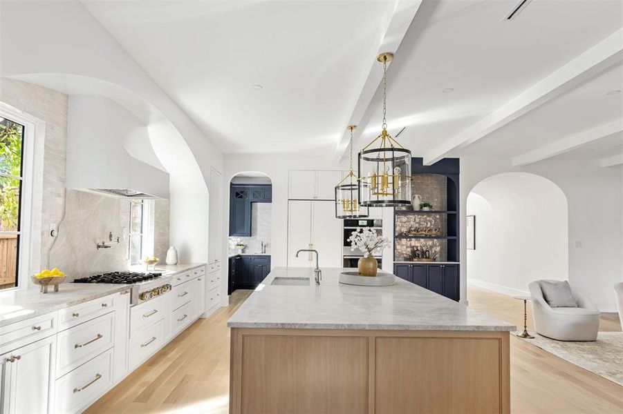 Kitchen featuring arched walkways, light stone countertops, an island with sink, beam ceiling, and white cabinets Kitchen featuring arched walkways, light stone countertops, an island with sink, beam ceiling, and white cabinets