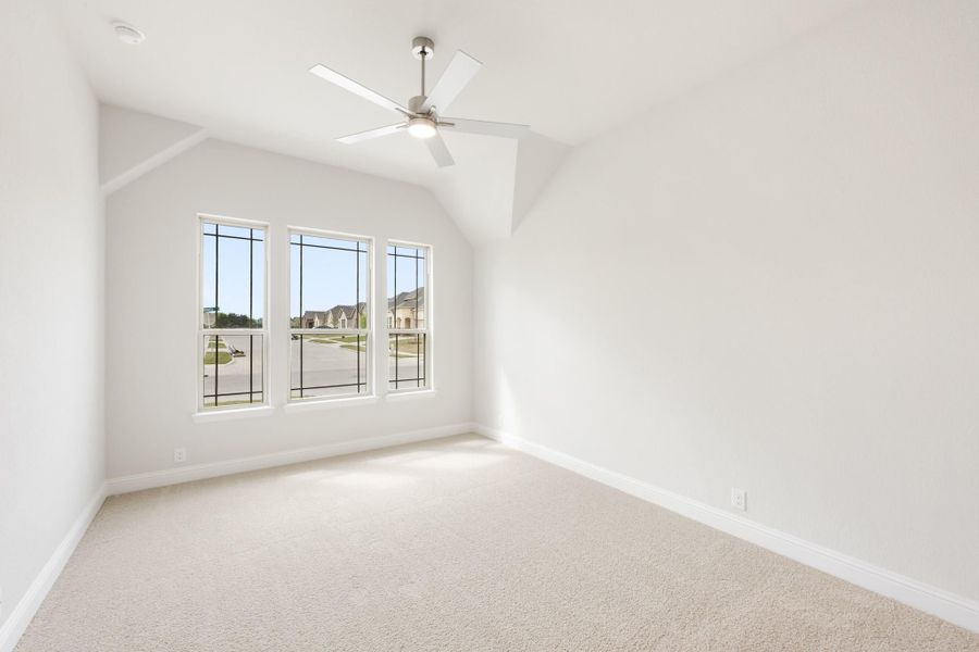 Empty bedroom with vaulted ceiling, ceiling fan, carpet flooring, and three windows overlooking a neighborhood street.