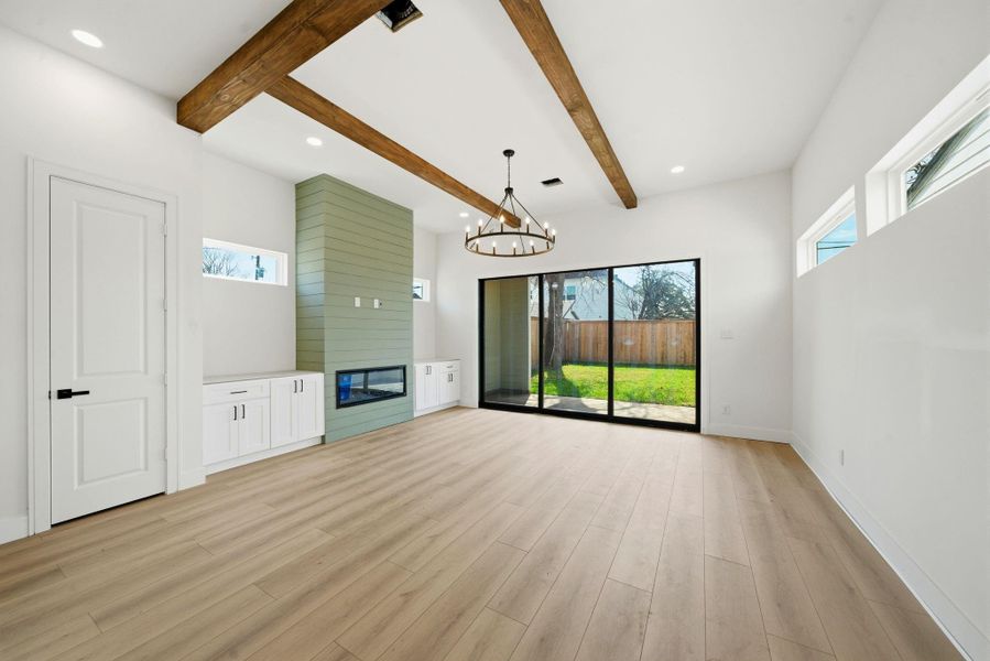 This spacious room features light wood flooring, exposed wooden ceiling beams, and a modern chandelier. It includes a built-in fireplace with cabinetry, large sliding glass doors leading to a backyard, and ample natural light from multiple windows.