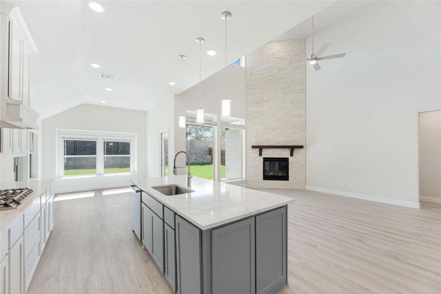 Open kitchen with gray island, white countertops, and vaulted ceiling open to living room with stone fireplace.