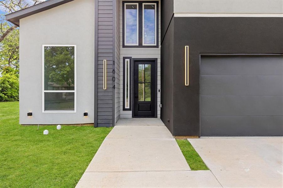 The property features a contemporary exterior with contrasting light and dark grey finishes, a prominent black entrance door with glass panels, and a dark grey garage door The property features a contemporary exterior with contrasting light and dark grey finishes, a prominent black entrance door with glass panels, and a dark grey garage door