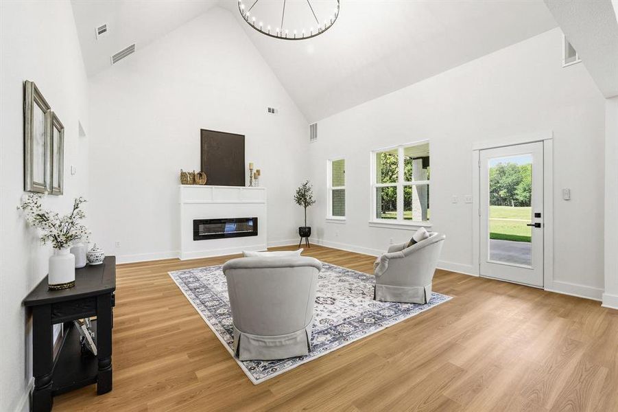 Living area featuring soaring vaulted ceilings, wood-finish flooring, a contemporary linear fireplace with a white mantel, and ample natural light from multiple windows and a glass-paneled door Living area featuring soaring vaulted ceilings, wood-finish flooring, a contemporary linear fireplace with a white mantel, and ample natural light from multiple windows and a glass-paneled door