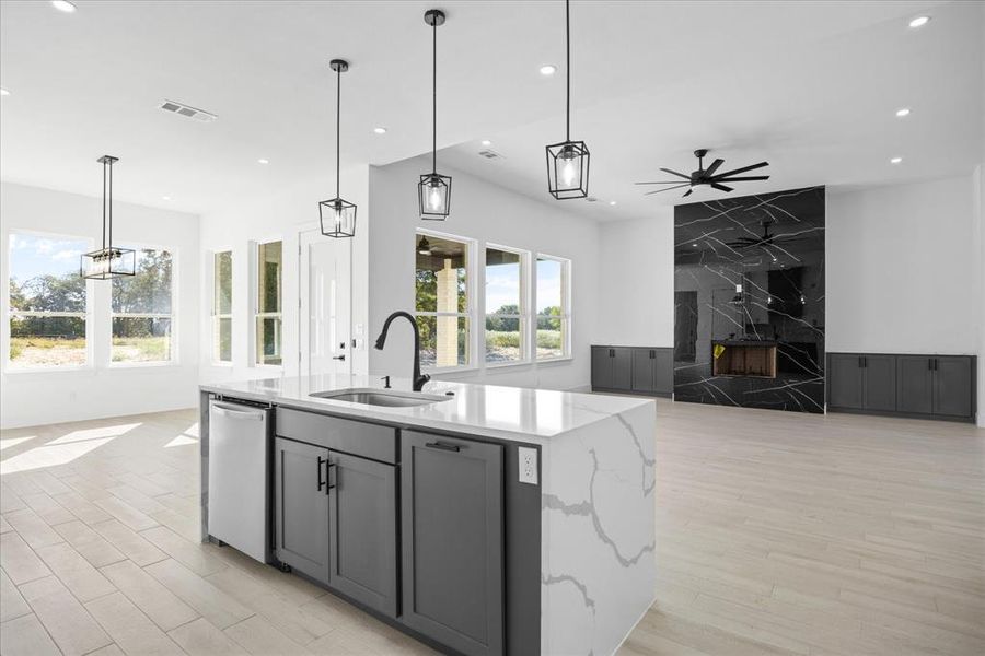 Spacious open-concept interior featuring light wood-finish flooring, a kitchen island with stone countertops and an undermount sink, a black and white accent wall with a built-in fireplace, and expansive windows