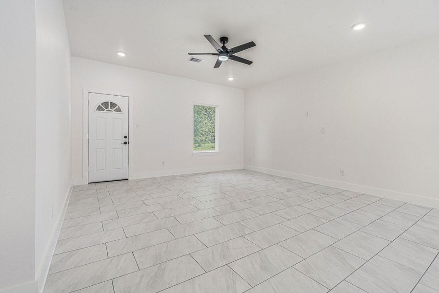 Bright and spacious room with modern tile flooring, a ceiling fan, recessed lighting, and a single window. The room features a white entry door and neutral walls, offering a clean and versatile space.