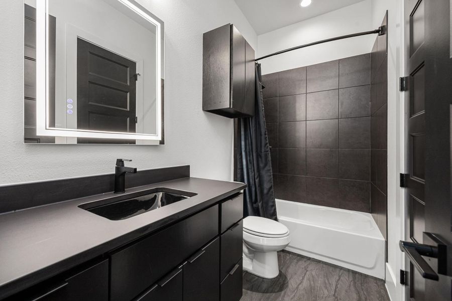 This modern secondary bathroom features sleek dark cabinetry and fixtures, an illuminated mirror, and a tiled shower-tub combo, providing a stylish and functional space. This modern secondary bathroom features sleek dark cabinetry and fixtures, an illuminated mirror, and a tiled shower-tub combo, providing a stylish and functional space.