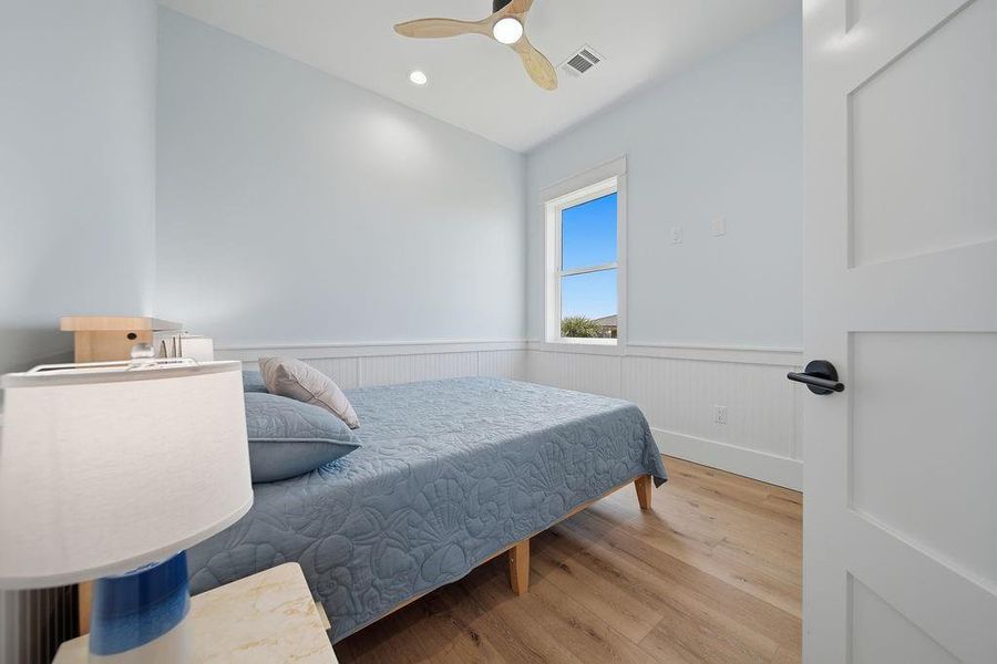 This cozy bedroom features light blue walls, wood flooring, and a ceiling fan. It includes a large window for natural light and a comfortable bed, creating a serene and inviting space.