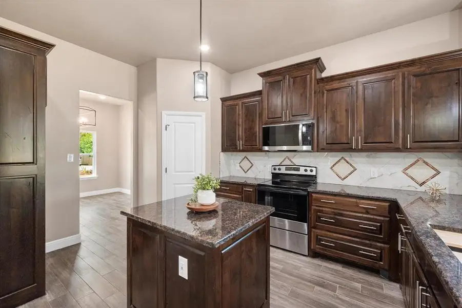 Kitchen featuring a central island with a granite countertop, knotty alder cabinets with hardware, stainless steel appliances, a decorative porcelain tile backsplash, and wood-finish flooring
