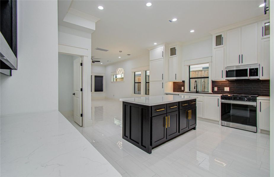 Home features a bright open-concept layout with sleek white walls and glossy tile flooring. The spacious kitchen includes a central island with dark cabinetry and gold accents, complementing the elegant wine rack and sleek design. The living area has large windows, a stylish light fixture, and access to an outdoor covered patio. A staircase leads to the upper level, enhancing the home's contemporary feel.