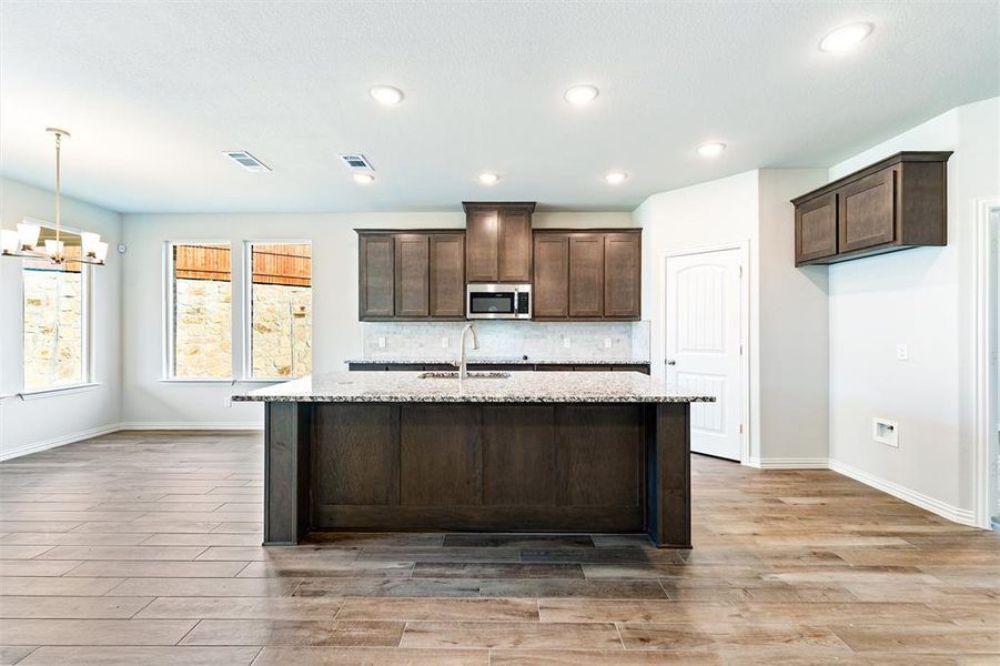 Kitchen with dark brown cabinets, light stone counters, a kitchen island with sink, a chandelier, and stainless steel microwave