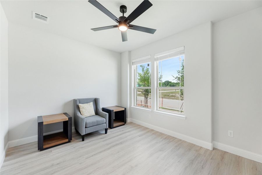 Sitting room with light wood floors, ceiling fan, gray accent chair, and two windows with outdoor views
